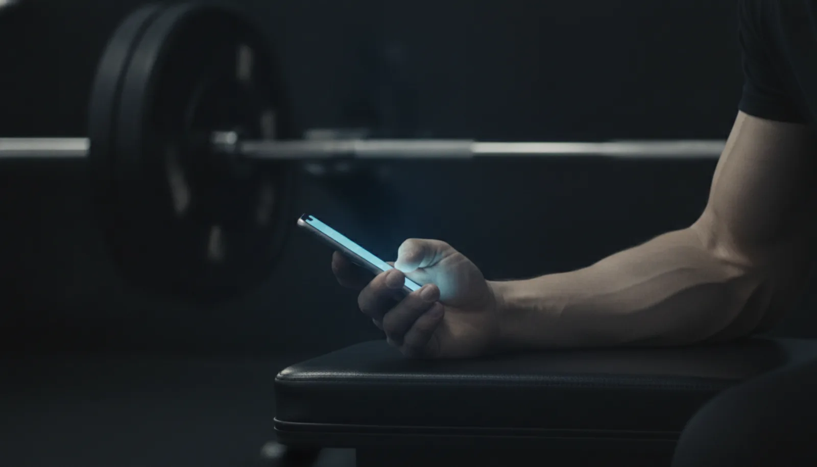 A man checking his phone on a gym bench beside a barbell