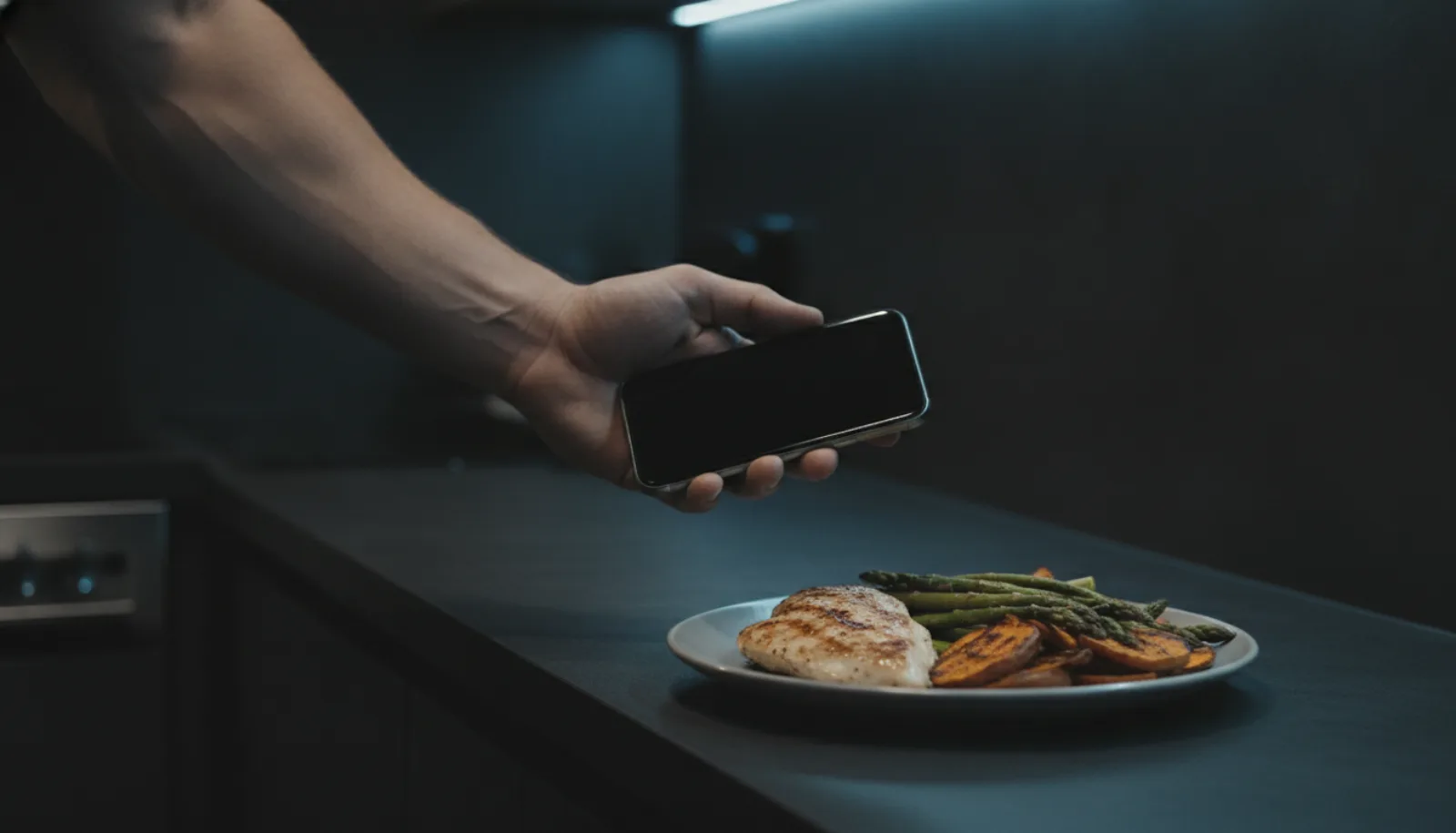 A man photographing a plate of food with his phone on a dark kitchen counter